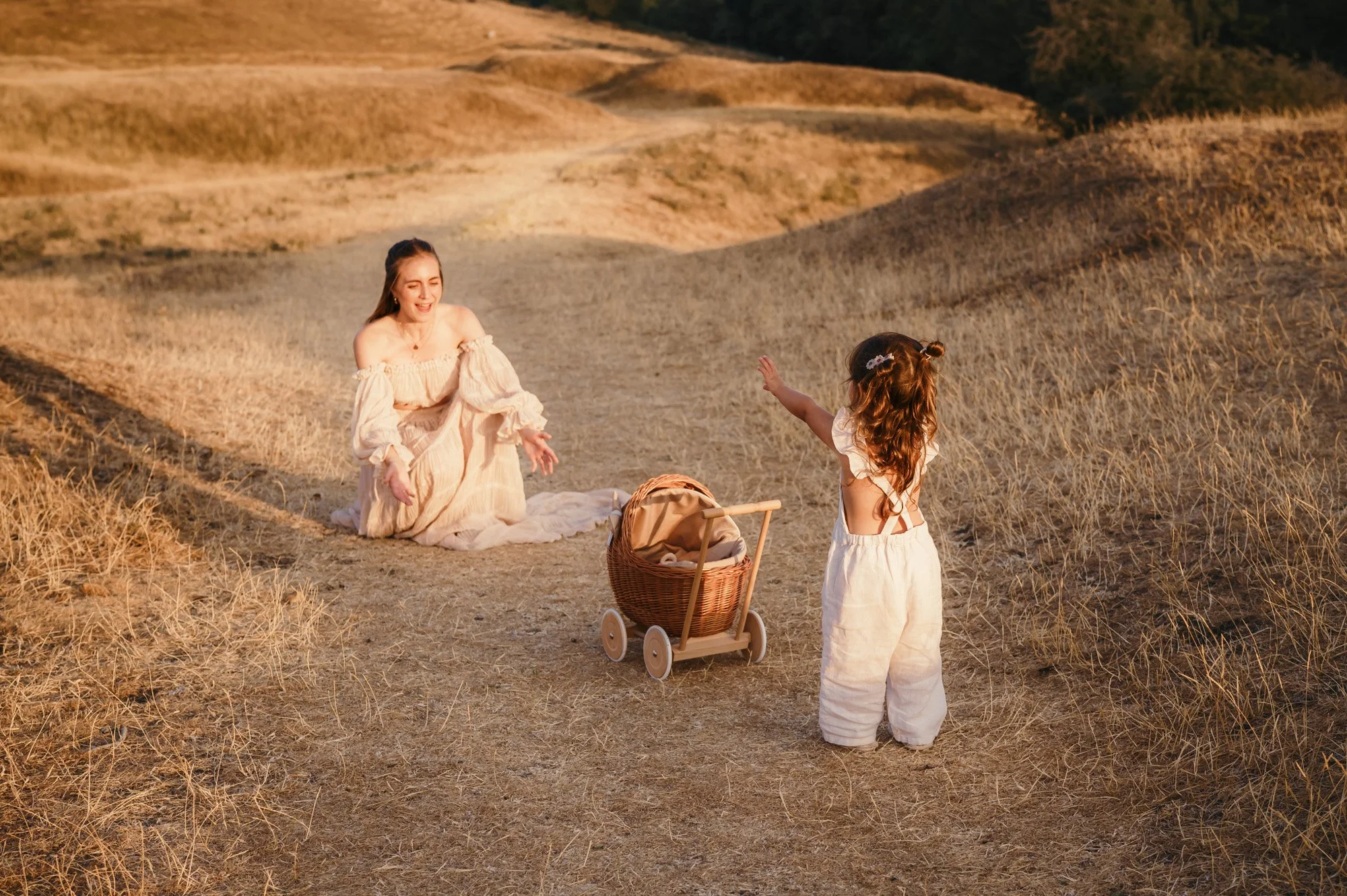 A woman in a cream-colored dress playing with a young girl in a white dress in a dry, hilly outdoor area during sunset. A wicker stroller is nearby.