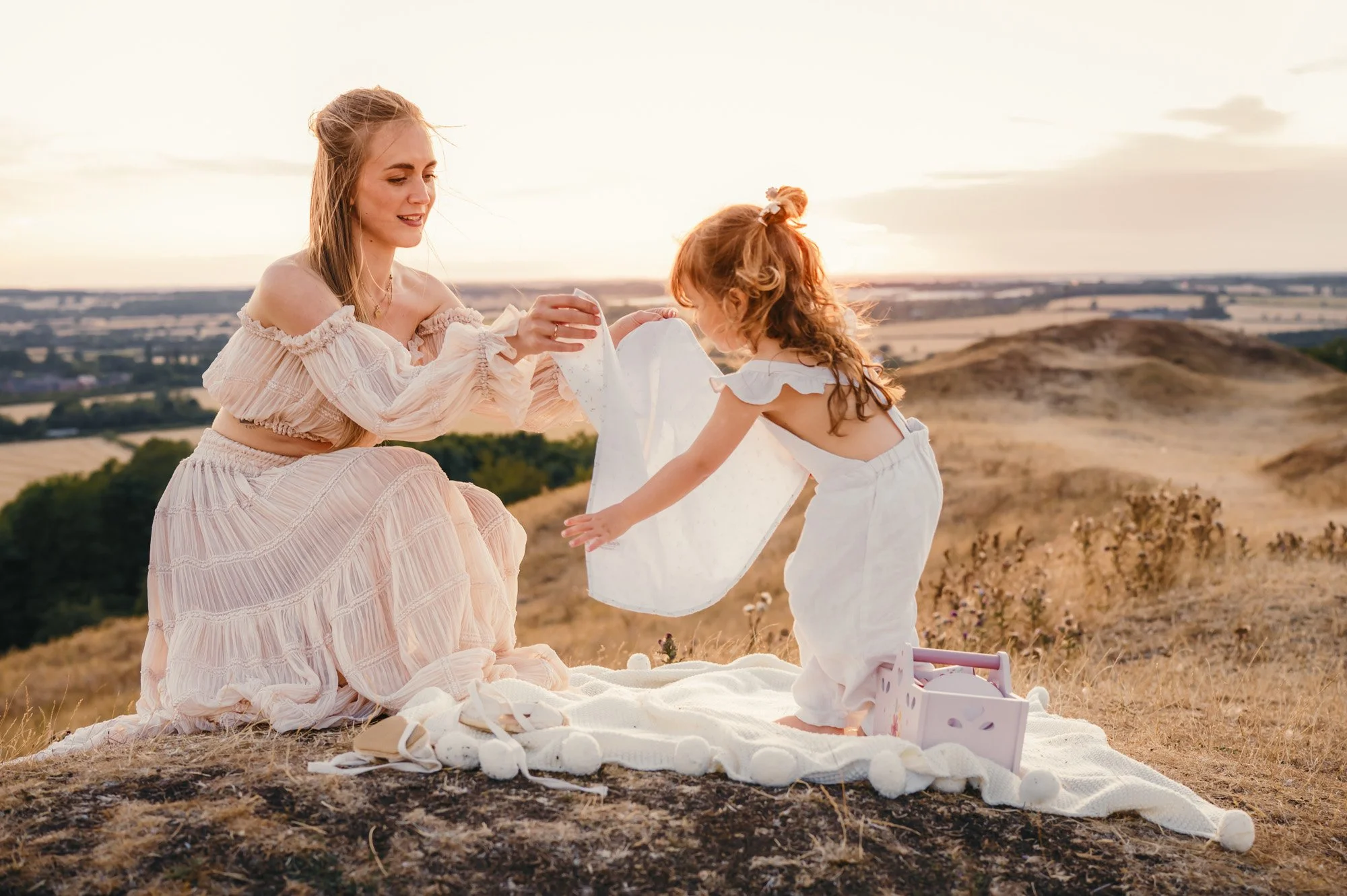 A woman and a young girl sharing a moment outdoors during sunset, with a scenic landscape in the background, on a blanket with a box of toys.