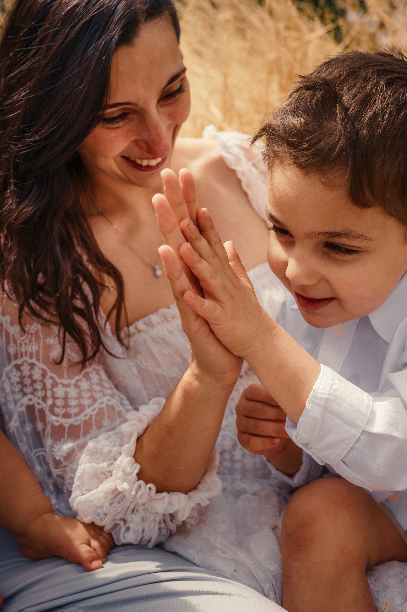 A woman and a young boy are engaged in a playful high-five. The woman has long, dark hair and is smiling, while the boy has short brown hair and is smiling in return. They are indoors, dressed in light-colored clothing, sitting close together and sharing a joyful moment.