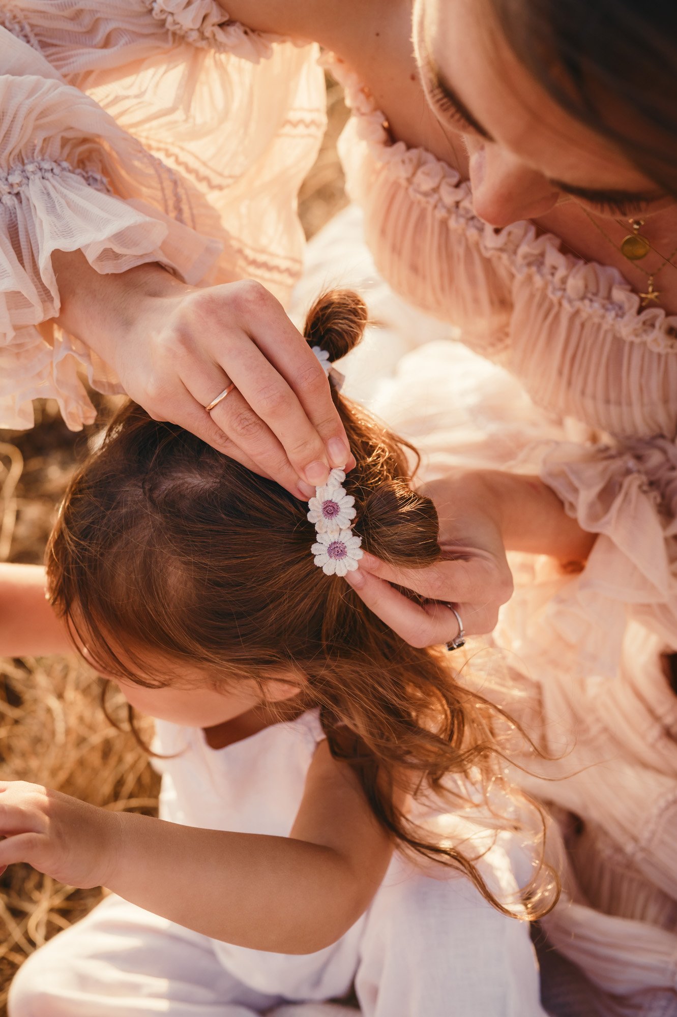 A woman with brown hair is placing a floral headband on a young girl with brown hair. The girl is wearing a white dress, and the woman is dressed in a light, ruffled, pink dress. They appear to be outdoors during sunset.