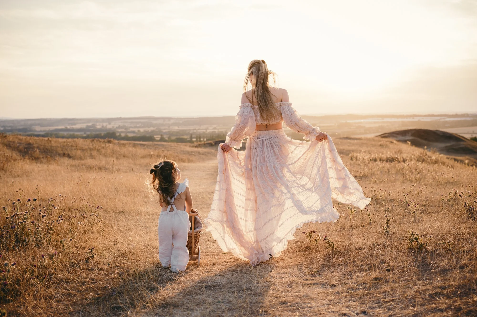 A woman and a young girl walking through a field at sunset. The woman holds her flowing dress while the girl carries a basket, both dressed in light, airy clothing.
