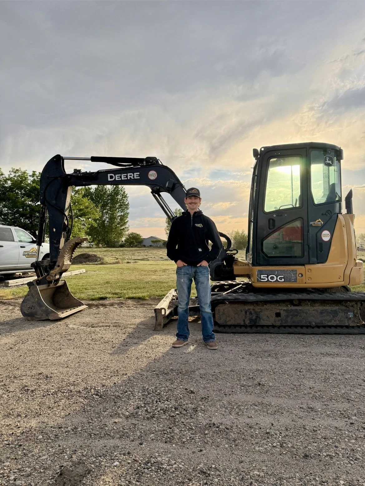 A young man wearing a black jacket, blue jeans, and a cap standing with hands in pockets in front of a yellow John Deere mini excavator at sunset. There is grass, trees, a white vehicle, and a small building in the background.