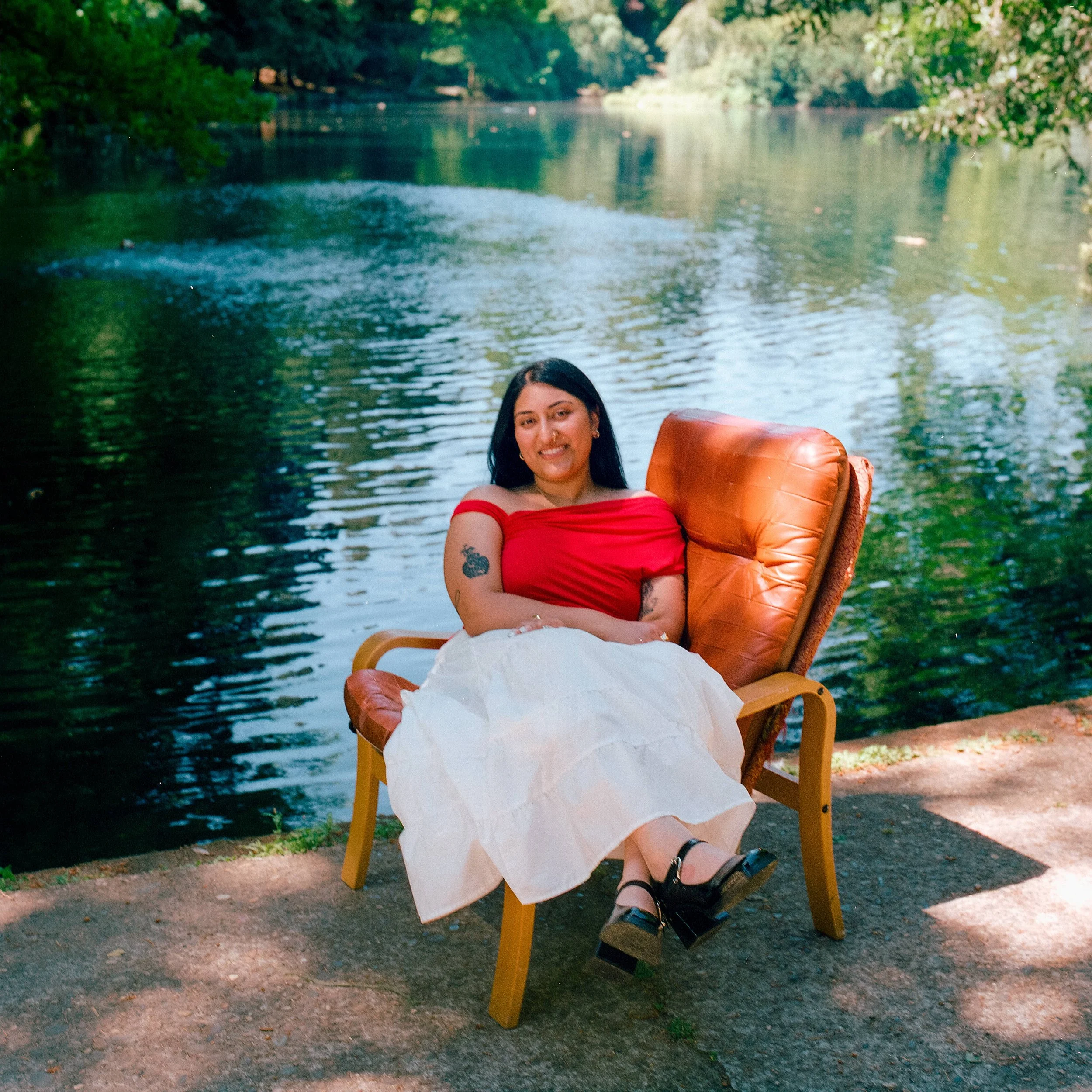 A woman sitting on a wooden chair by a river, holding an orange cushion, surrounded by green trees and water, wearing a red top and white skirt.