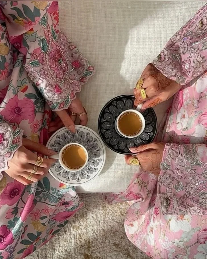 Two women wearing colorful, embroidered dresses, each holding a cup of tea or coffee on decorative saucers, visible from a top view.