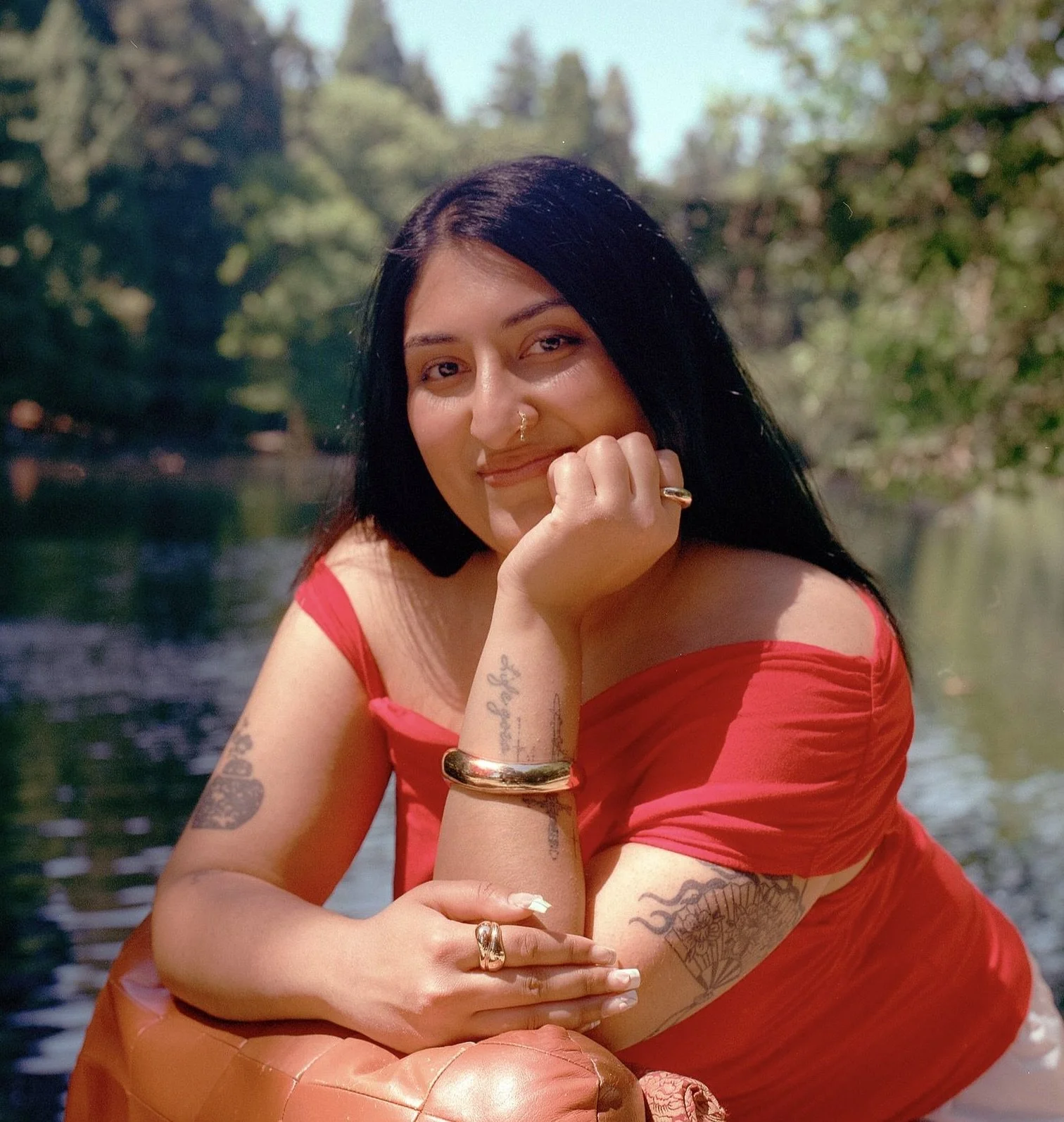 A woman with black hair, tattoos, and rings, wearing a red off-shoulder dress, sitting outdoors near water with a background of trees, smiling and resting her chin on her hand.