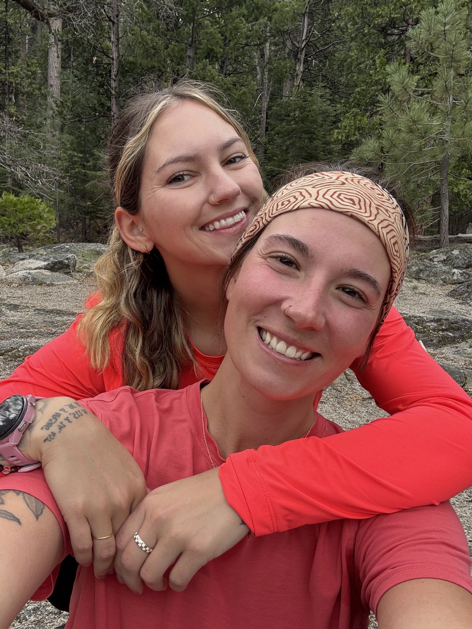 Two women smiling, taking a selfie outdoors in a forested area with trees and rocks.