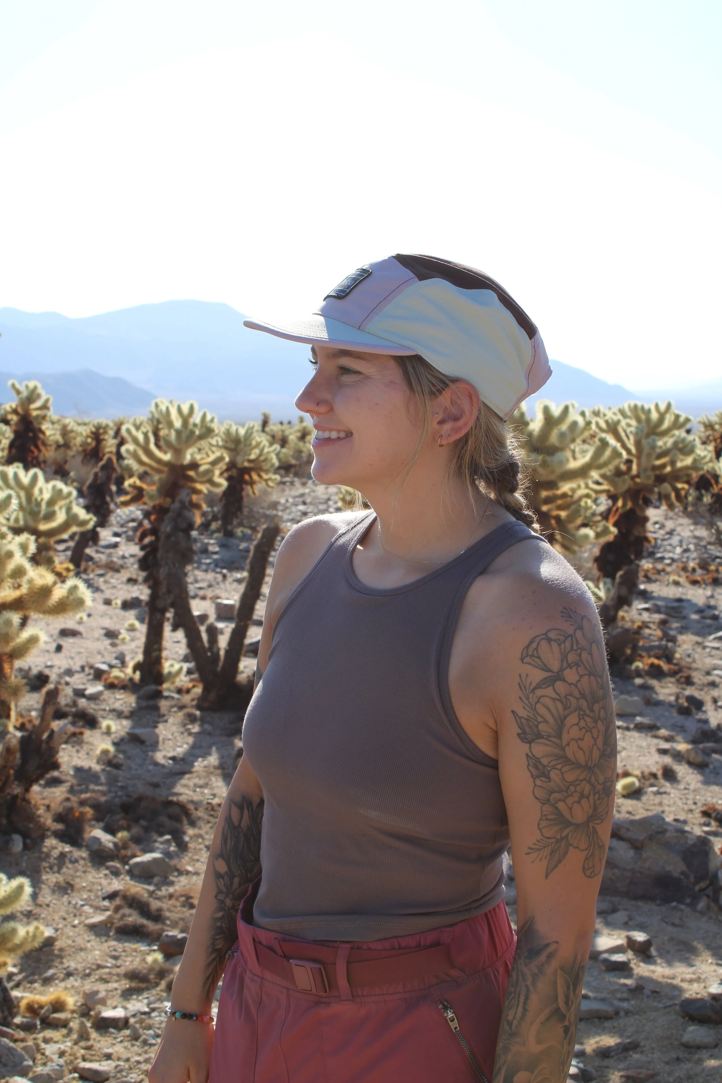 A woman with tattoos on her arm, wearing a gray tank top, pink pants, and a hat, smiling while standing in a desert with cacti and mountains in the background.