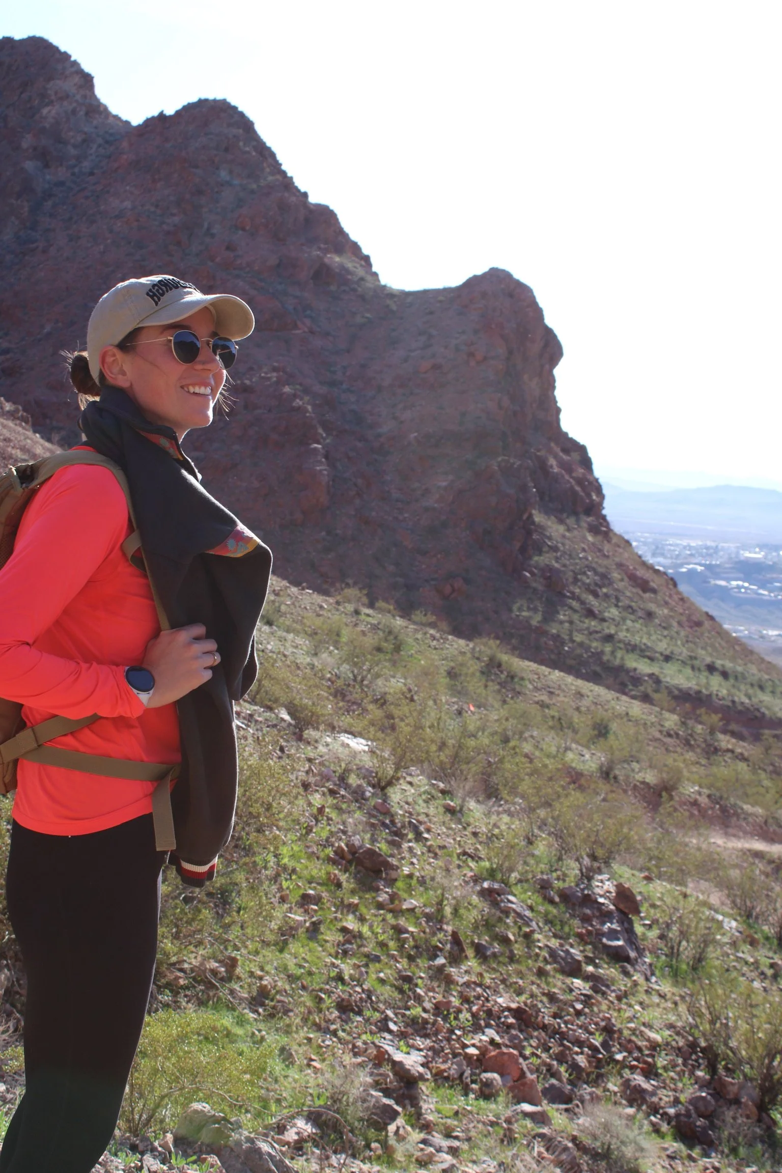 A woman hiking on a rocky trail in a mountainous area with red rock formations, wearing sunglasses, a beige cap, a pink jacket, and a backpack.