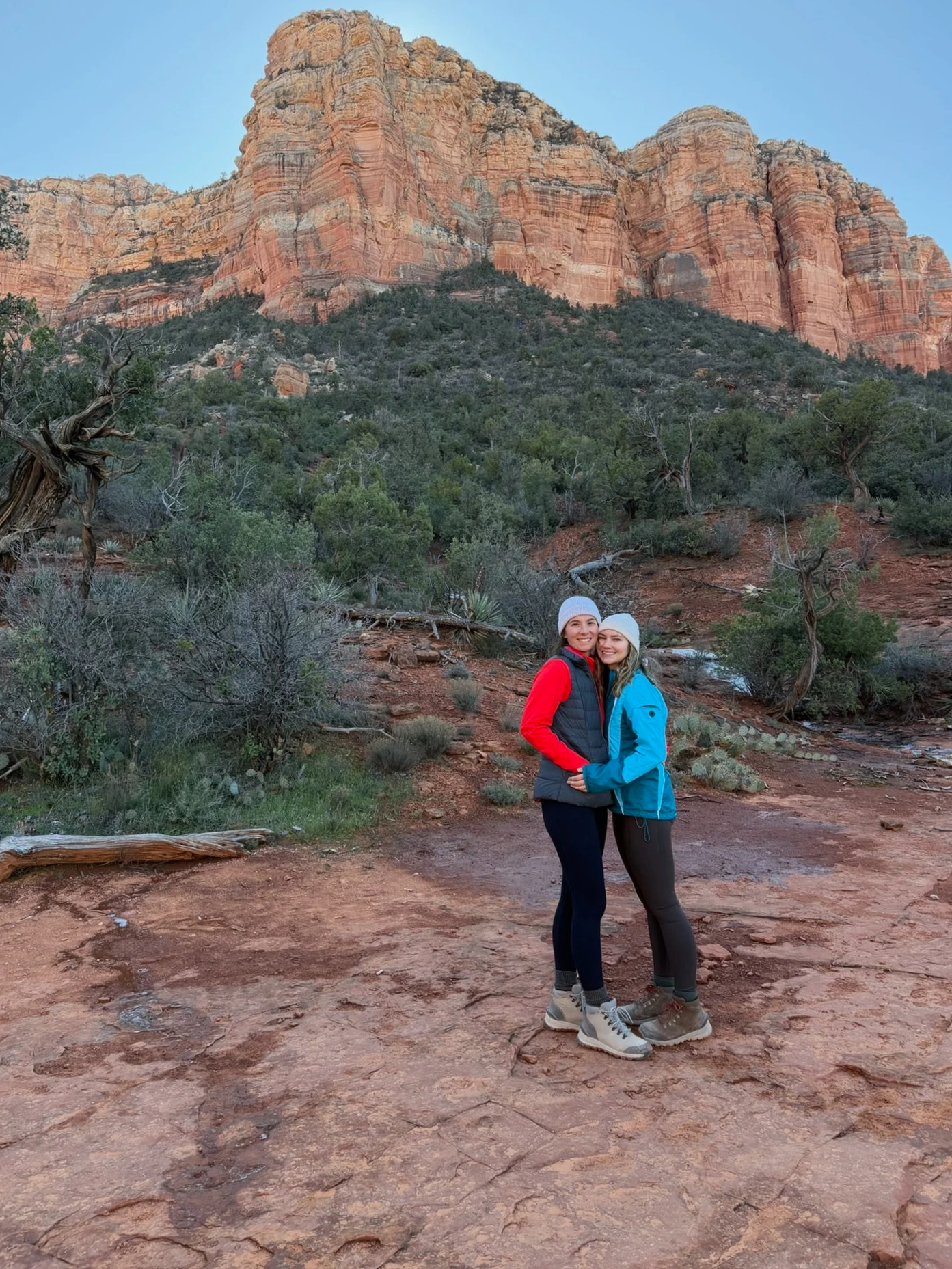 Two women in outdoor winter clothing standing together on a dirt trail in a red rock canyon with a large cliff in the background, smiling at the camera.
