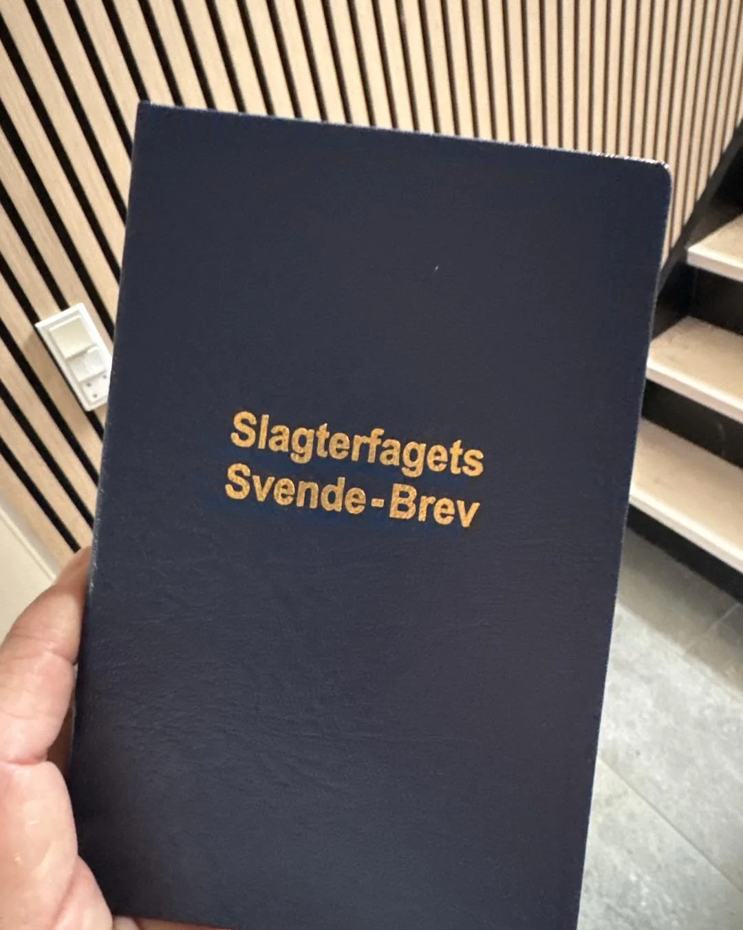 Billedet viser en hånd, der holder en blå mappe med gul tekst, der siger 'Slagerfagets Svende-Brev'. I baggrunden er der en væg med trælister og et stikkontakt.