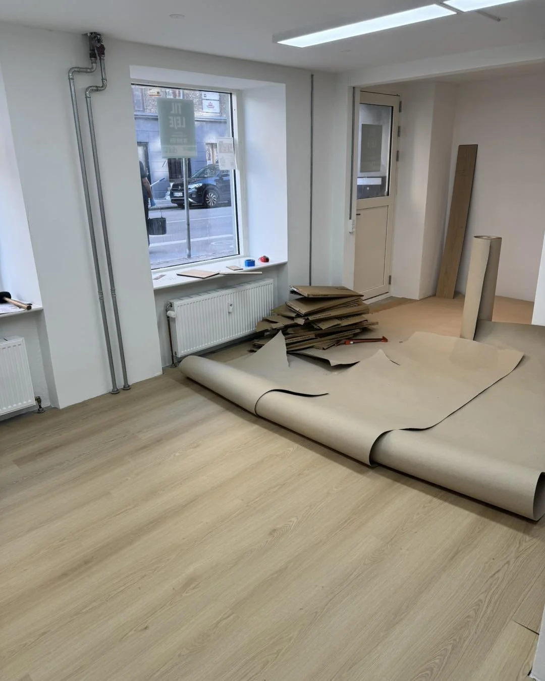 Indendørs rum under renovation med papirruller, værktøj og vindue, væggene er hvide og gulvet er lyst træ.