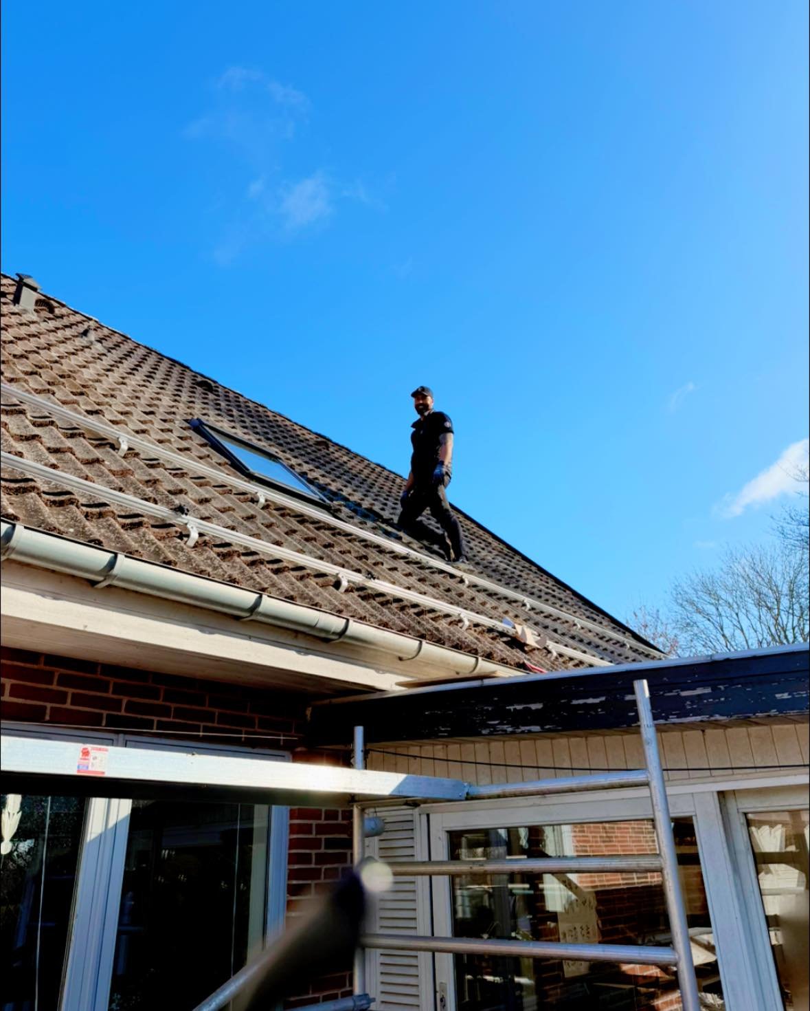 En mand arbejder på et hustag med tagrender og skifferbelægning under en klar blå himmel.