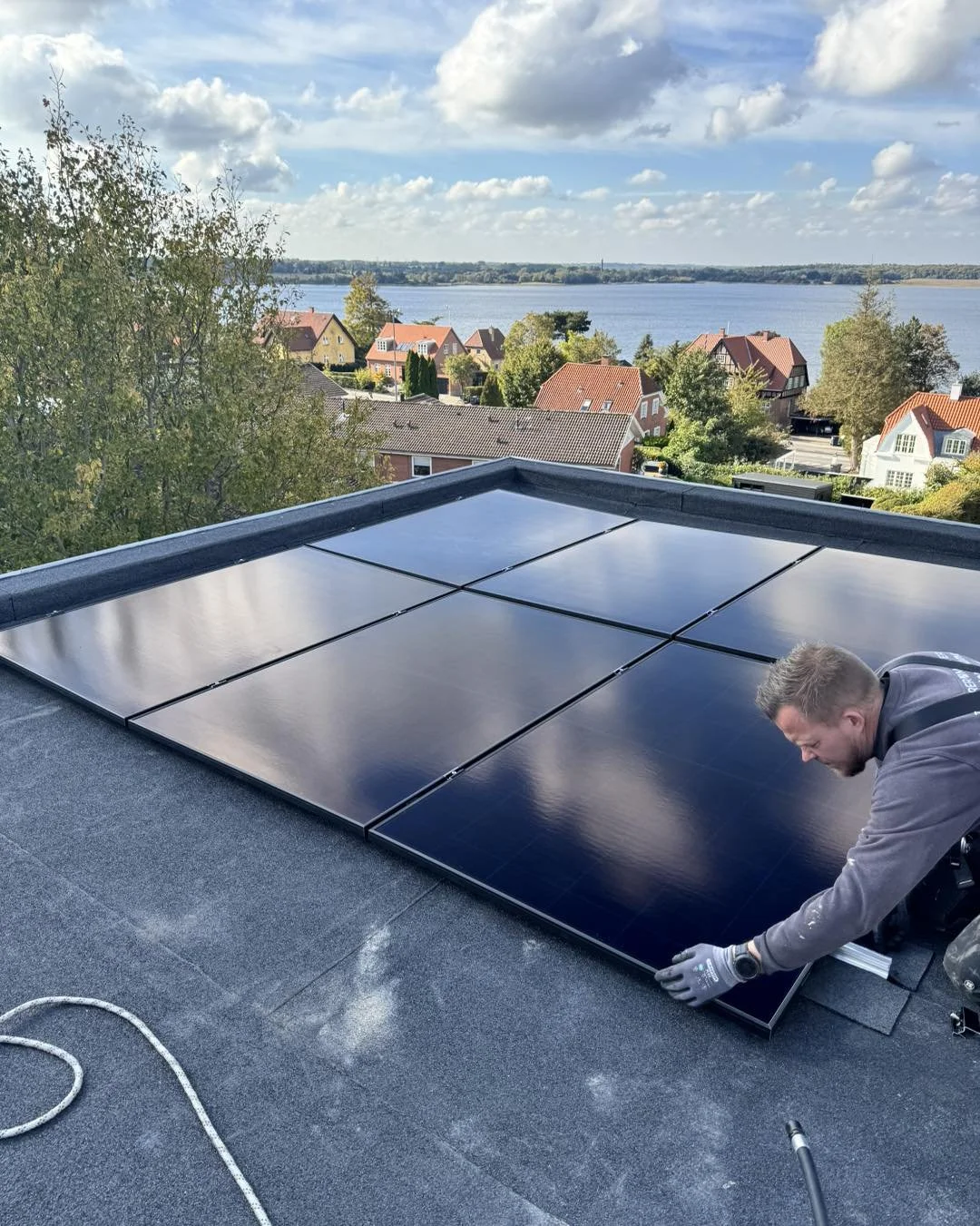 En mand installerer solceller på taget af en bygning med udsigt over en sø og huse i baggrunden.