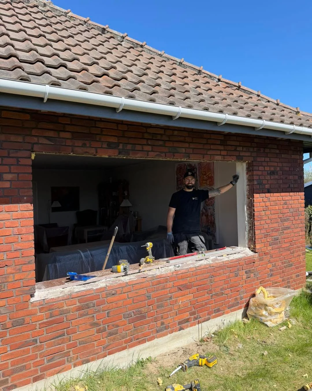 En mand står foran et stort åbent vindue, der er under renovering, i en rødbrygget væg. Han smiler og peger mod væggen. Der er værktøj og materialer på vindueskarmen, og en grøn have står udenfor.