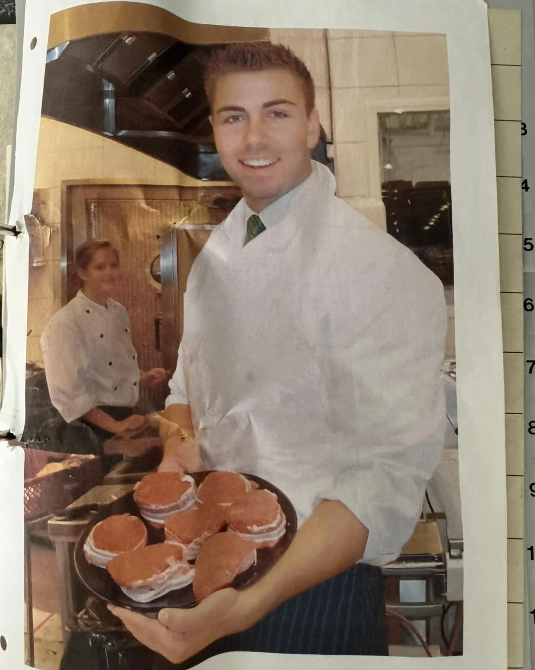 En mand, der smiler og bærer en skål med burgere, med en kvinde i kokkeuniform i baggrunden i et køkken.