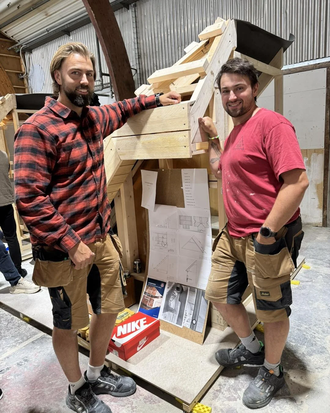 To mænd står ved en udsigt lavet af trælægter, smiler og ser stolte ud. Der er arkitektplaner og en Nike skotøjsæske på gulvet.
