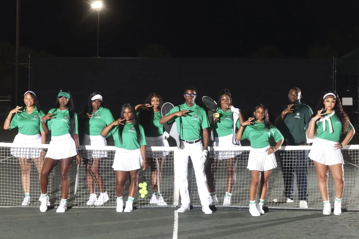 A group of ten young women and two men posing on a tennis court at night. The women are wearing green shirts, white skirts, and tennis shoes, holding tennis rackets, with some making peace signs. The men are dressed in black and green shirts and white pants, standing among the women. There is a tennis net in front of them and stadium lights illuminating the court.