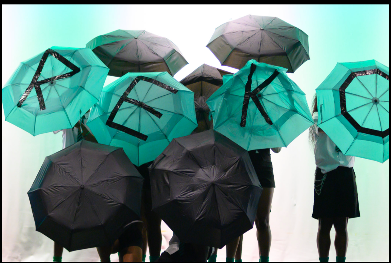 Group of people standing beneath teal and black umbrellas, with some umbrellas displaying the letters R, E, K, O, and O.