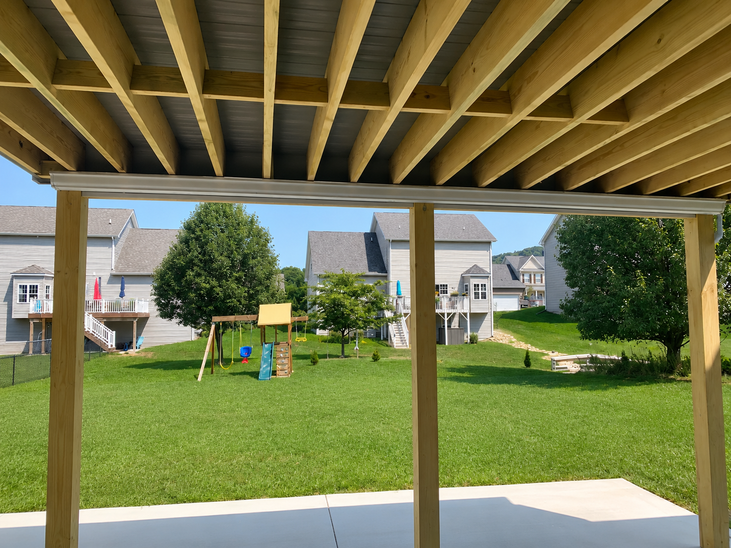 View of a backyard with green lawn, children's playset, and trees, as seen from under a wooden porch with supports.