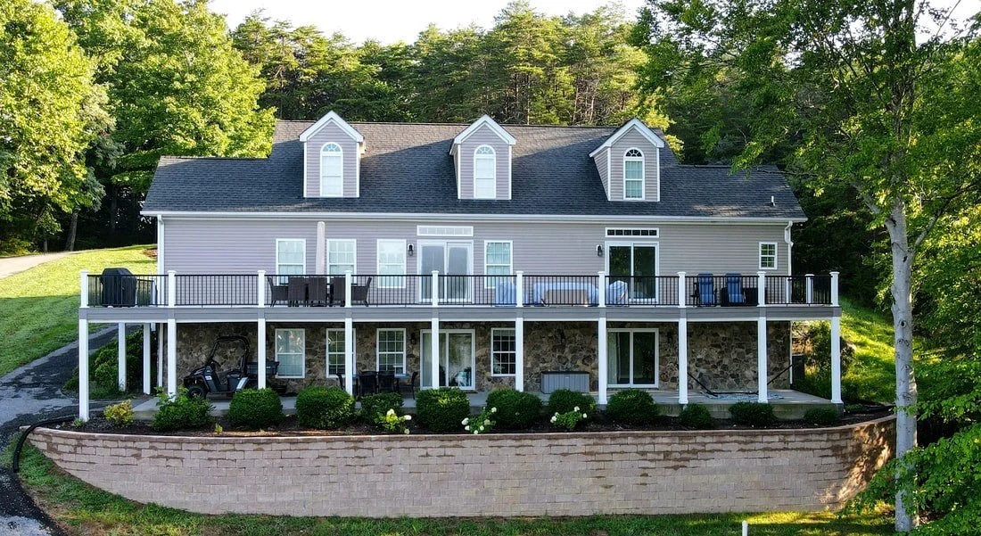 Two-story house with a stone foundation, beige siding, and a dark roof, surrounded by trees, featuring a large outdoor deck with porch furniture and a golf cart underneath.