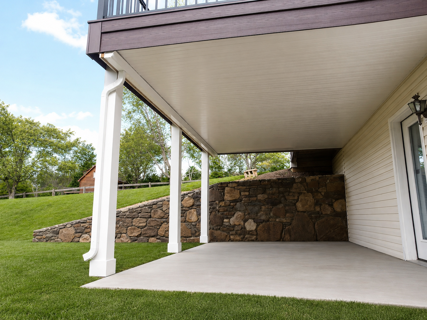 Back porch area of a house with white support beams, beige siding, a stone retaining wall, and a grassy yard with trees in the background.