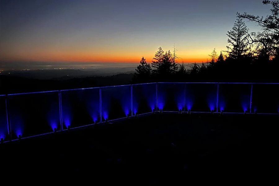 A sunset view from a scenic overlook with a blue-lit railing and silhouetted trees.