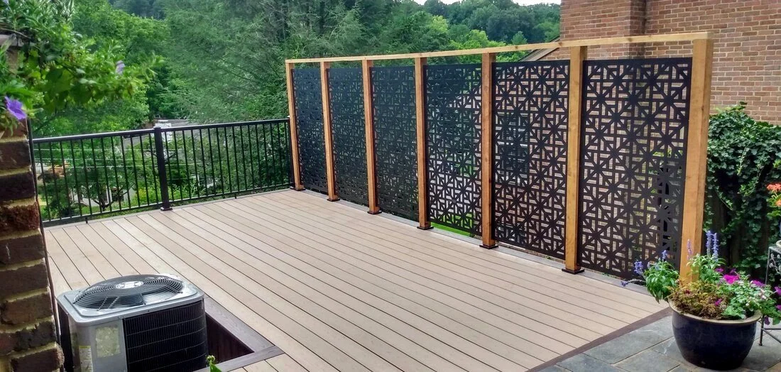 Newly built backyard deck with black decorative panel fencing, an air conditioning unit, potted plants, and a view of trees.