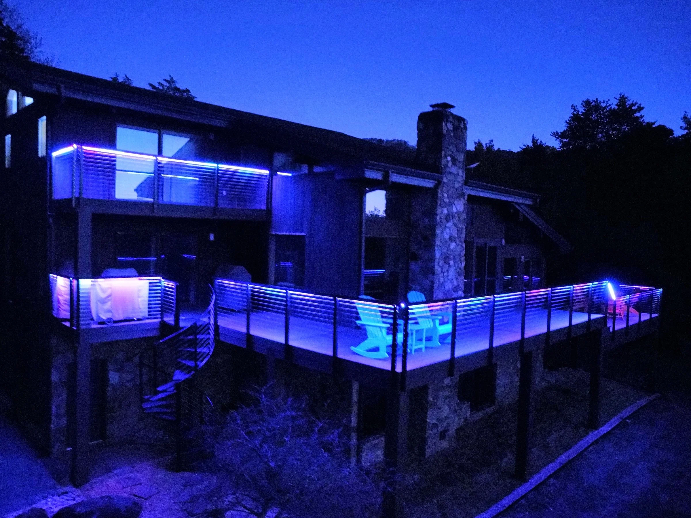 A house at night illuminated with blue and purple LED lights on the balcony and railing, with a stone chimney and trees in the background.