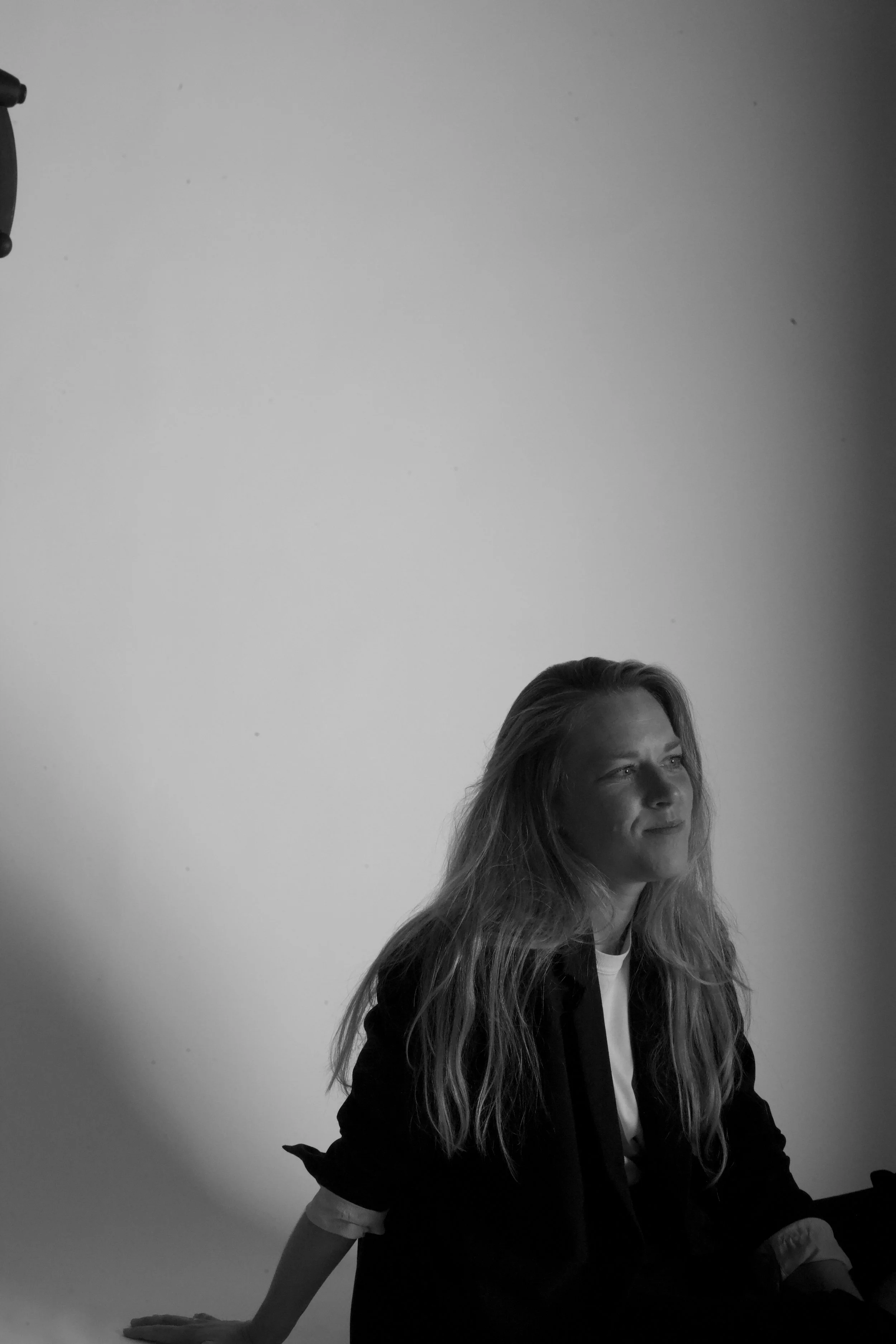 Een woman met lang haar zit aan een tafel, op de achtergrond een witte muur en een plafond met een lamp.