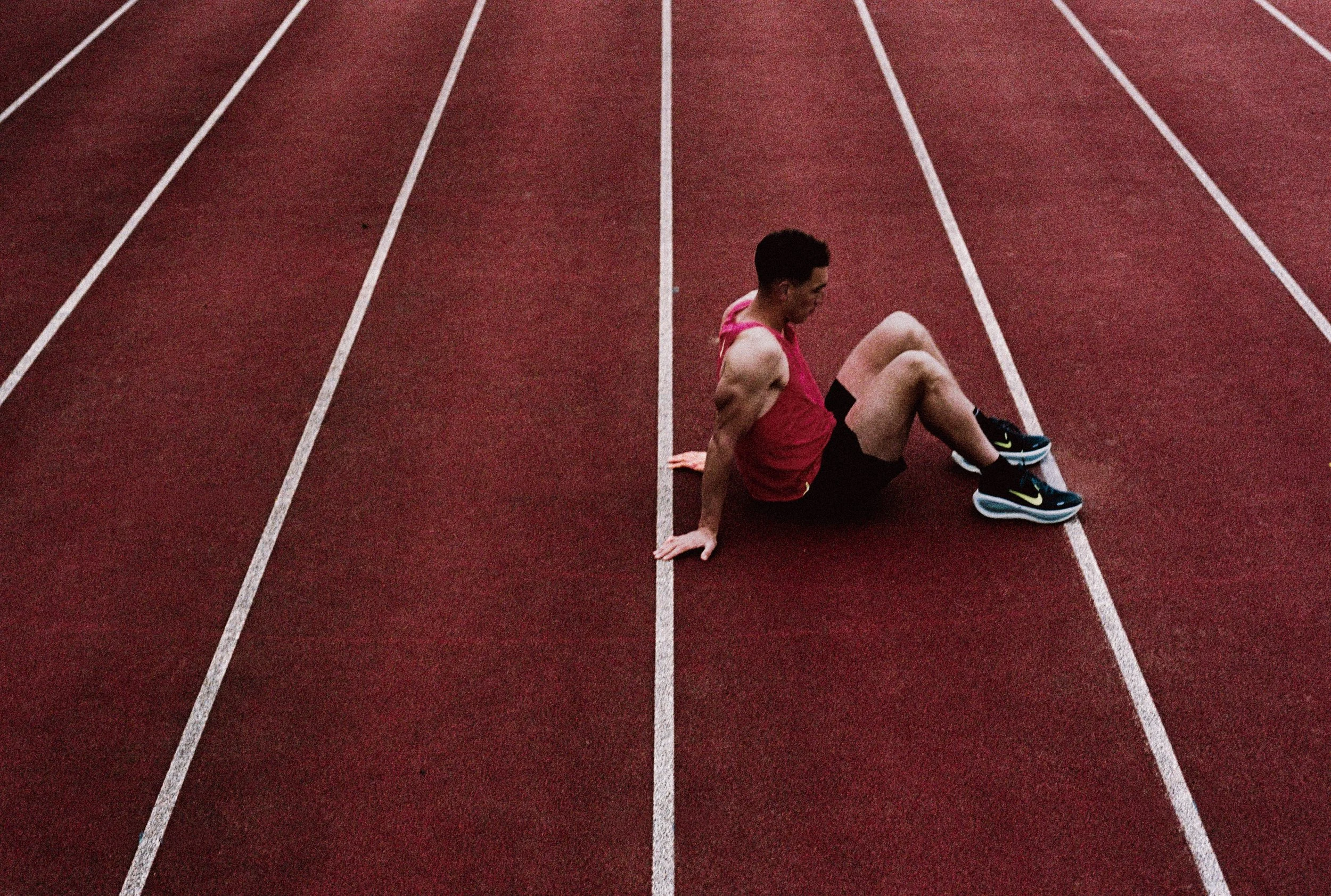 Een man in sportkleding zit op een atletiekbaan, zithoek met witte lijnen en een rood oppervlak.