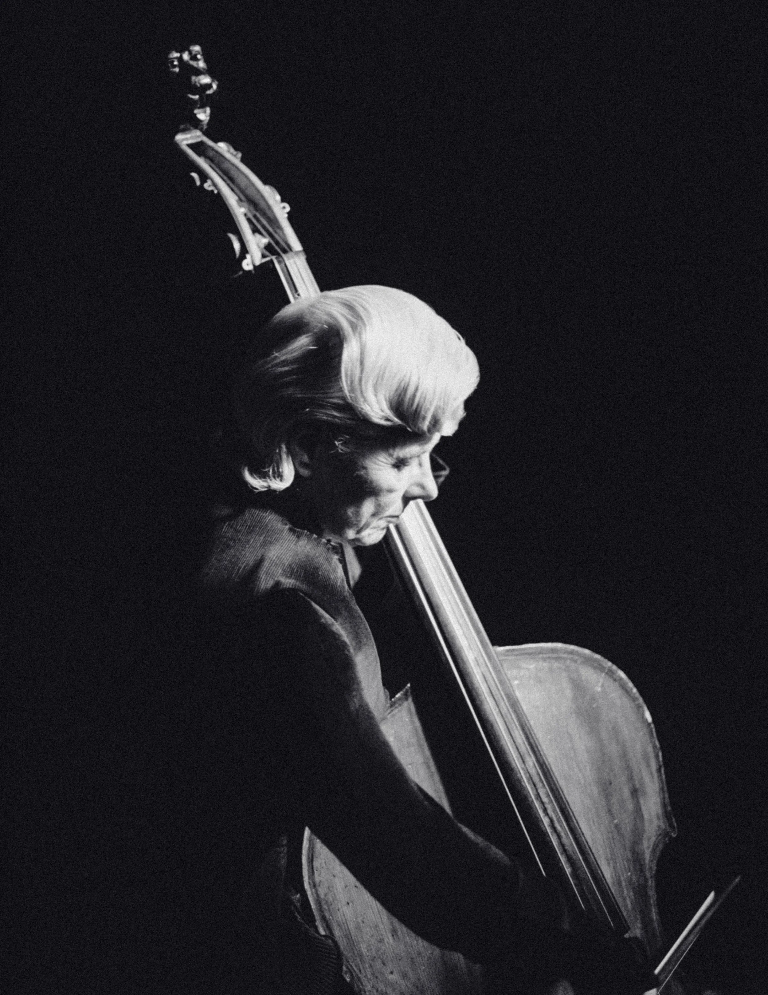 Zwart-wit foto van een vrouw die op een cello speelt. Ze heeft grijs, golvend haar en is geconcentreerd op haar spel.