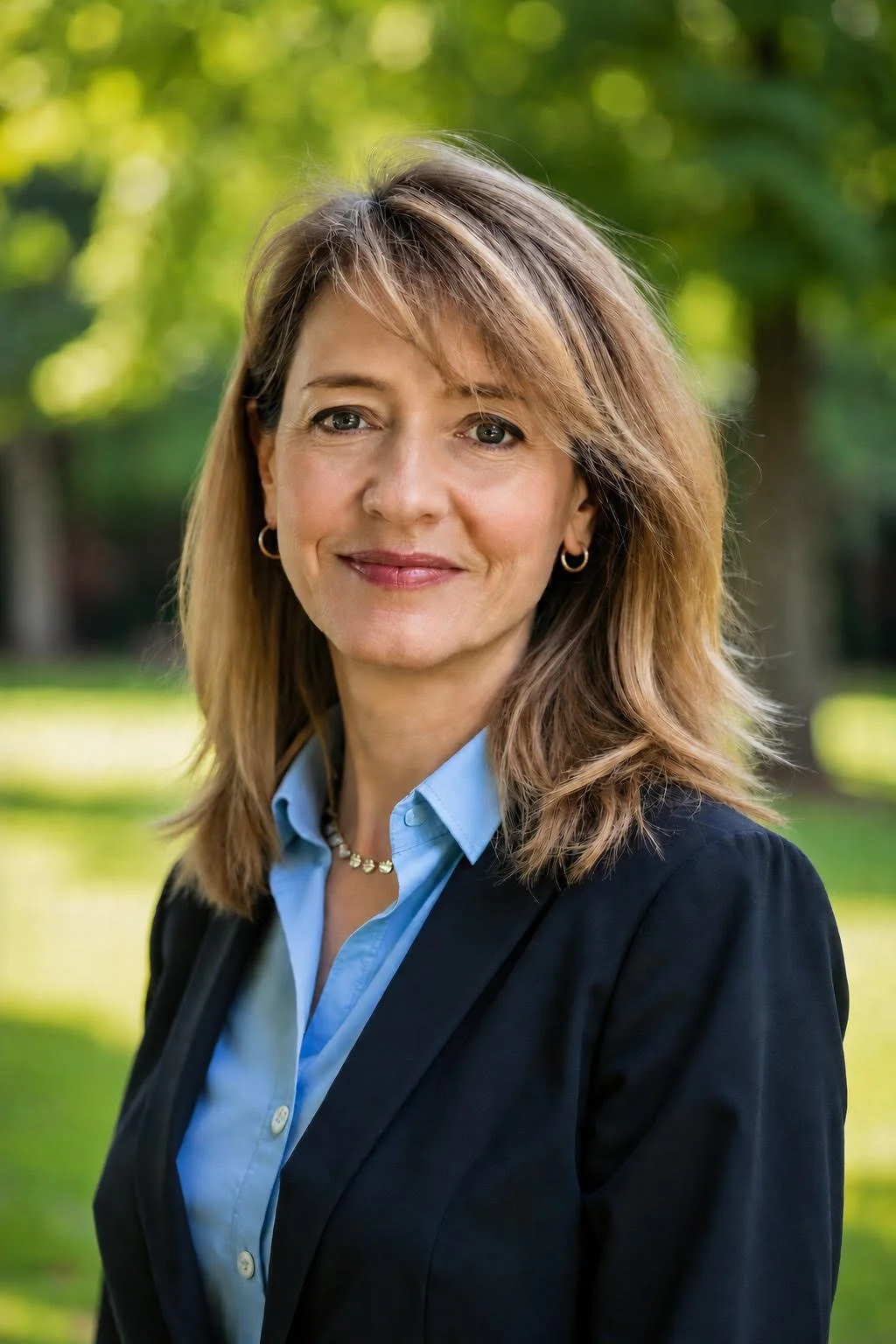 A woman with shoulder-length light brown hair, wearing a black blazer and a light blue shirt, standing outdoors with a blurred green park background.