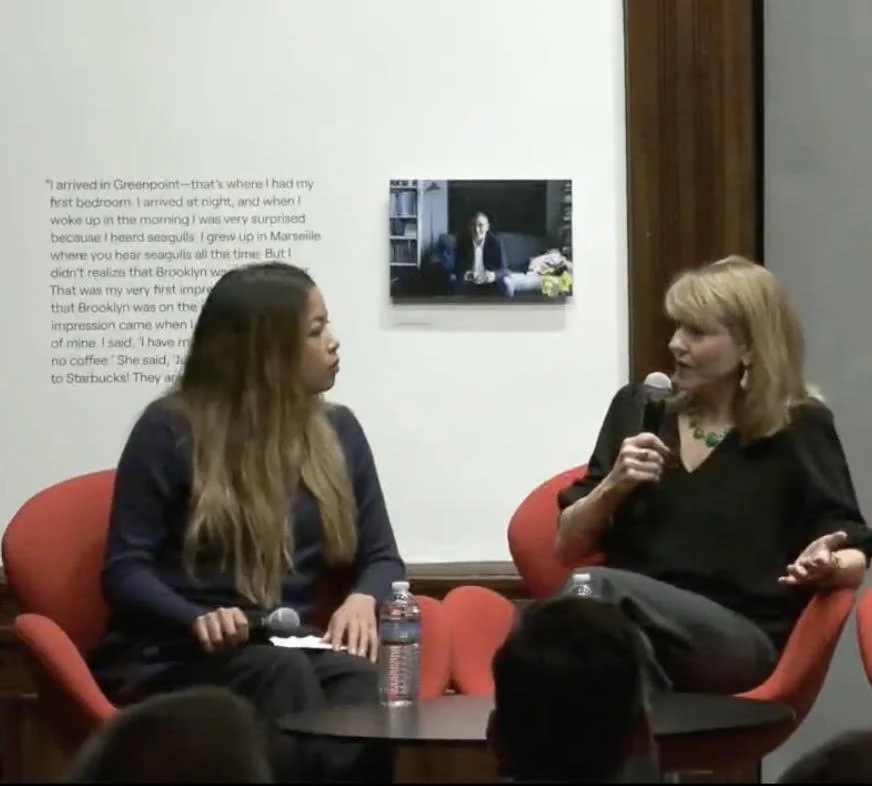 Two women are sitting on orange chairs in conversation. The woman on the right is holding a microphone, and the woman on the left has a water bottle and a notepad. A small table is between them. A wall with a quote and a small mounted TV showing a wo