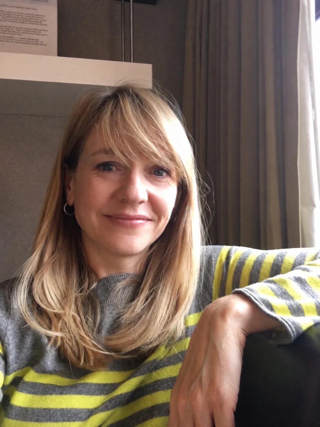 A woman with blonde hair, wearing a grey and yellow striped shirt, sitting indoors near a window with light-colored curtains, smiling at the camera.