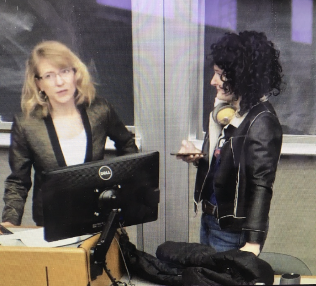 Two women in a classroom or office, one with blonde hair and glasses standing behind a desk with a computer, and the other with curly black hair and headphones around her neck looking at her phone.