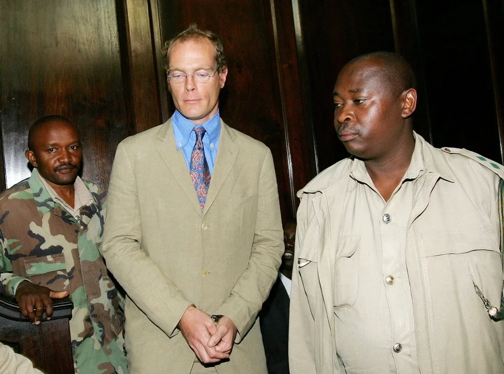 Three men, one in camouflage military uniform, one in khaki attire, and one in a beige suit jacket, standing indoors against a wooden wall, with serious expressions.