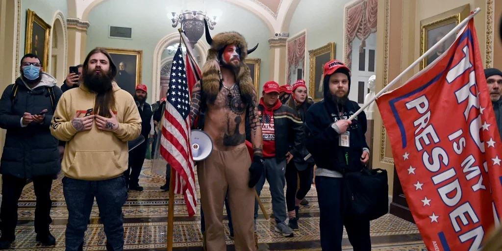 Group of people, some wearing Trump hats and holding a Trump 2020 flag, in an ornate room with gold framed paintings and a high ceiling.