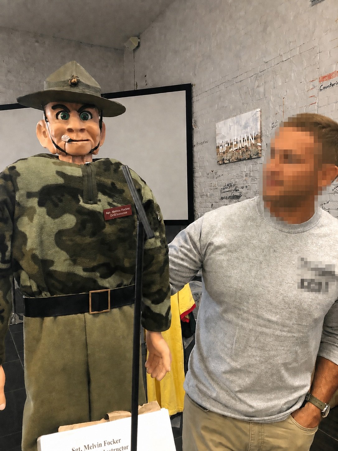 A person standing next to a large military mascot or statue dressed in camouflage and wearing a hat, with a badge that reads 'Sgt. Melvin Focker Drill Instructor,' while the person with blurred face is wearing a light-colored shirt.