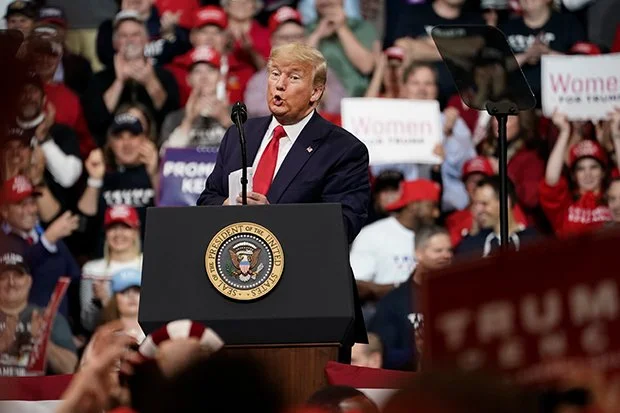 Former President Donald Trump speaking at a rally, standing behind a podium with the presidential seal, in front of a crowd holding signs and wearing red hats.