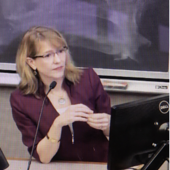 A woman with shoulder-length blonde hair, wearing glasses and a maroon blazer, sitting in front of a computer with a microphone, possibly giving a presentation or lecture in a classroom or conference setting.
