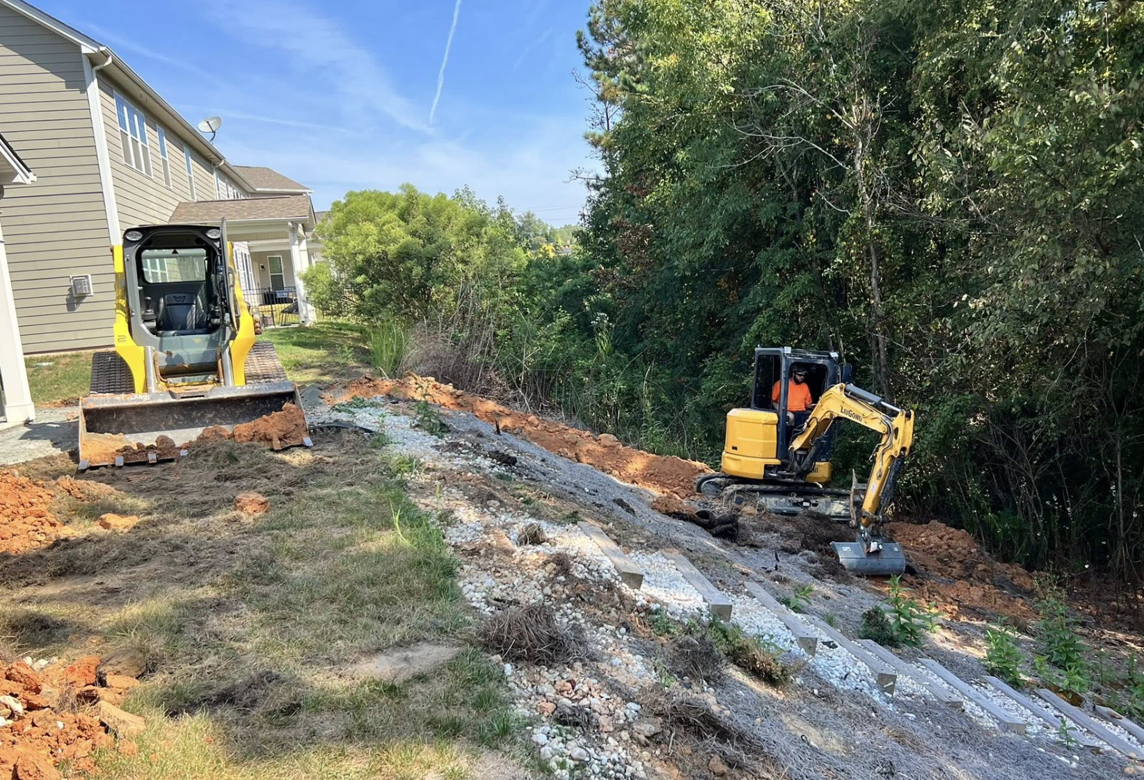Construction workers operating small excavators to build a pathway or retaining wall near a residential house. The area has been excavated with soil and gravel laid out.