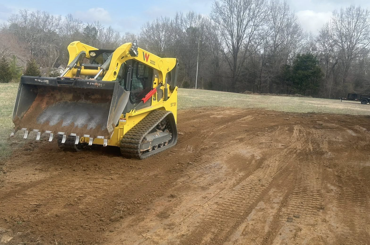 A yellow compact track loader with an open front bucket, working on a dirt lot, with trees and a trailer in the background.