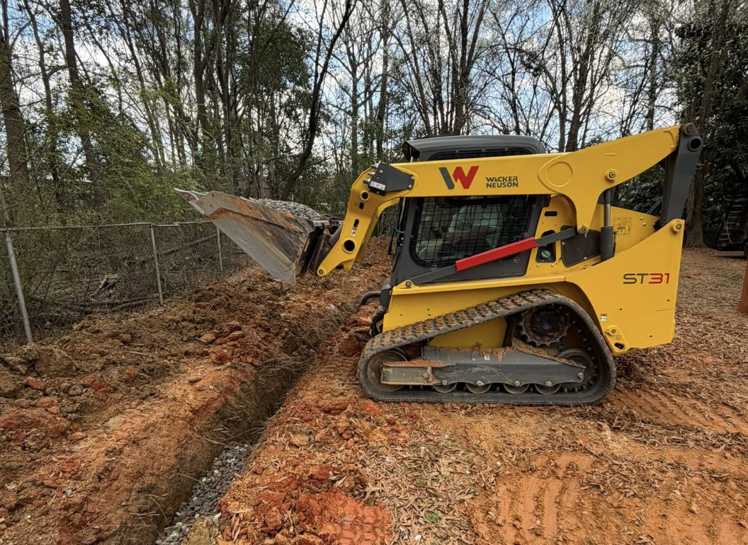 A yellow compact excavator with tracks, labeled Wacker Neuson, digging a narrow trench in a dirt area near trees and a chain-link fence.
