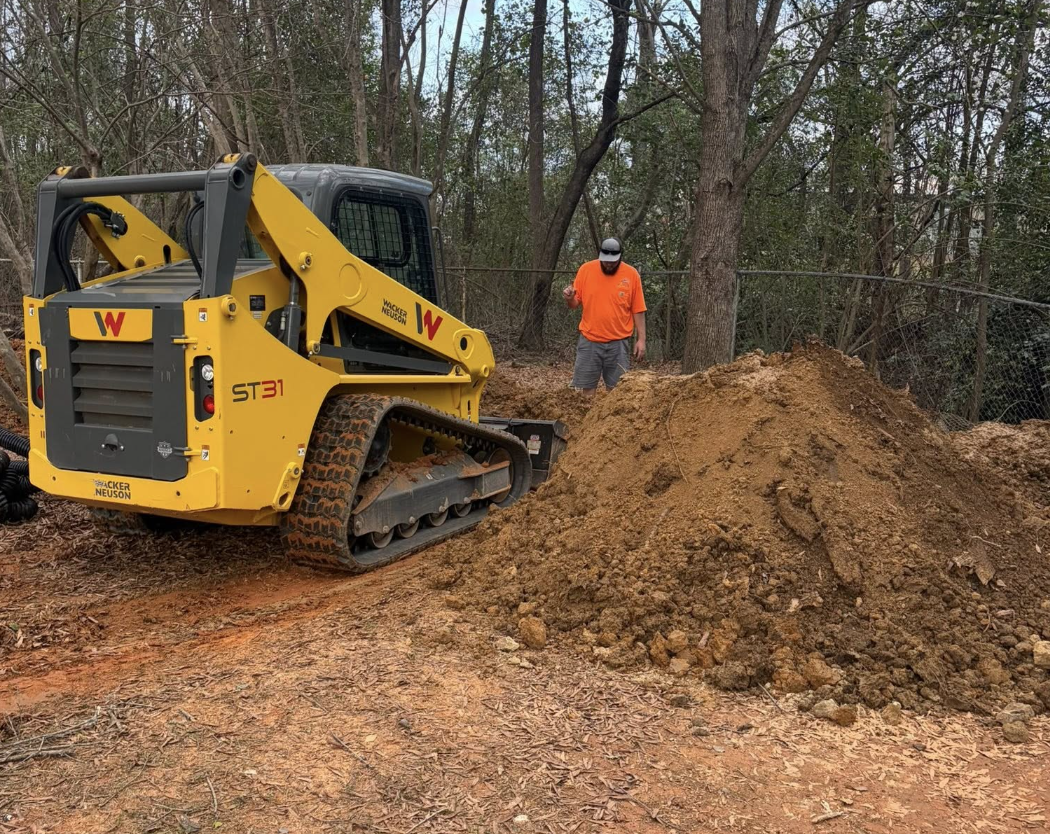 A construction worker in an orange shirt and hard hat standing next to a yellow compact track loader on a dirt trail with a pile of soil, surrounded by trees and a wire fence.