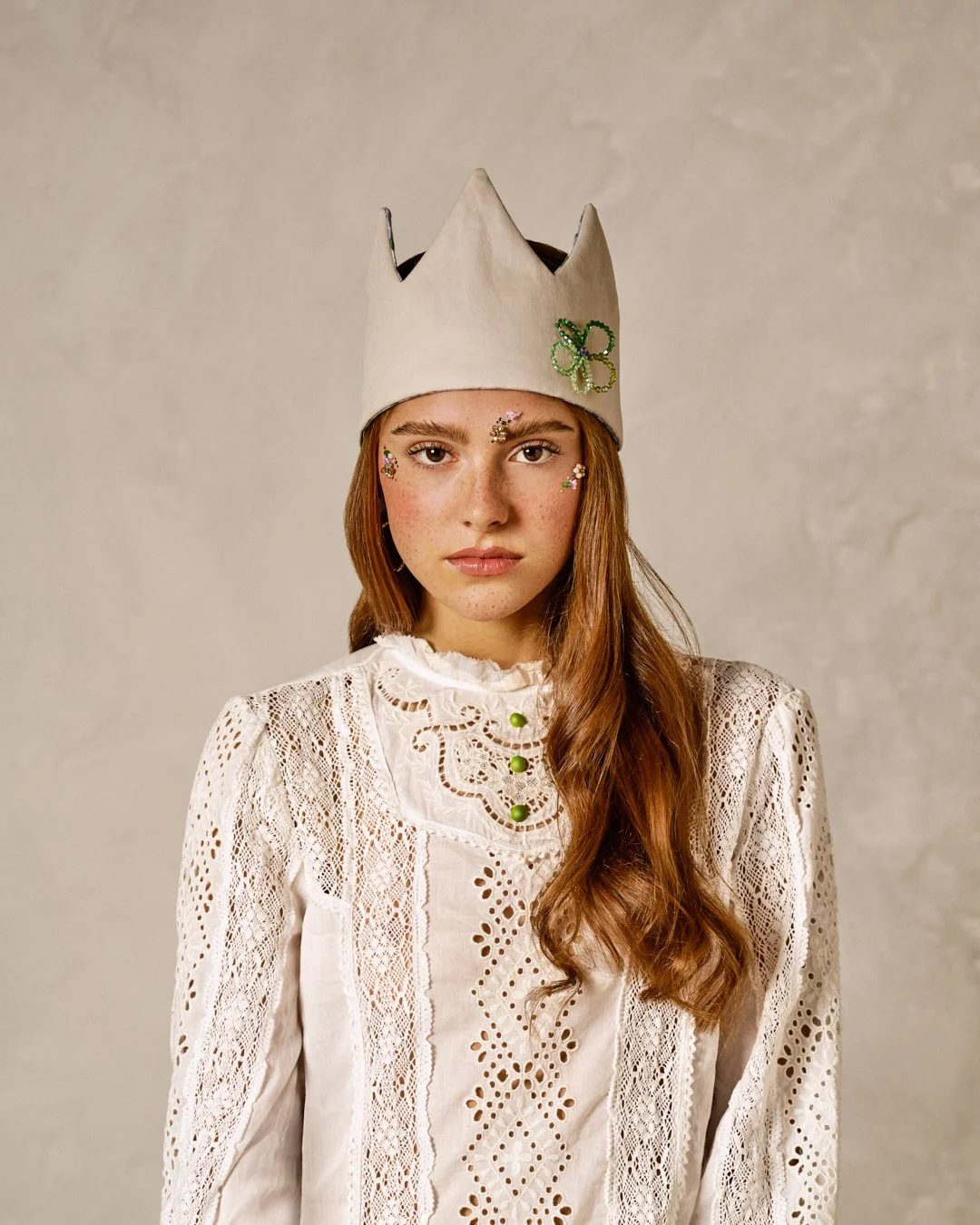 Jeune femme avec un chapeau en papier, décorée d'une feuille verte en perles, portant une robe blanche en dentelle, avec des petits autocollants en forme de fleurs sur le visage et le cou.