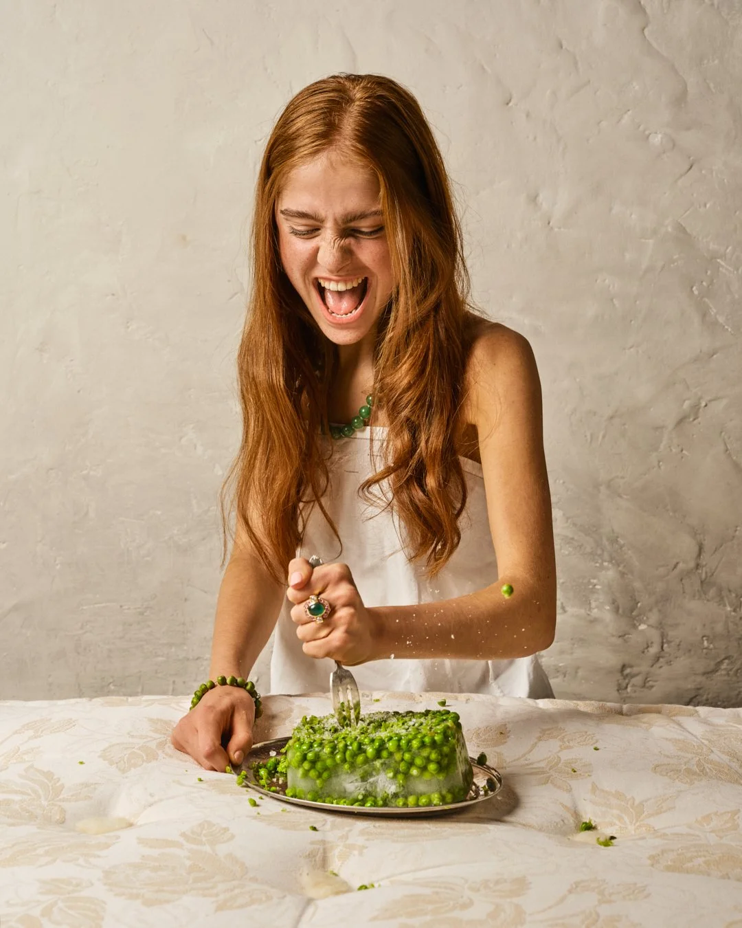 Une femme avec des cheveux roux, souriante, coupe un gâteau aux pois verts, habillée en blanc avec des bijoux verts, devant un mur beige