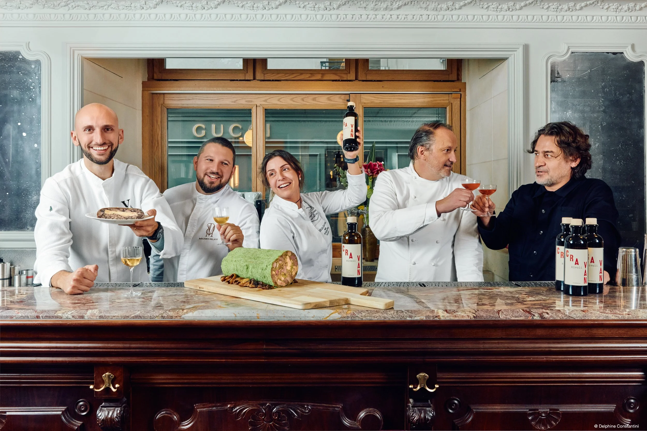Groupe de cinq personnes en tenue de cuisine derrière un comptoir en marbre, tenant des verres à vin et des bouteilles de vin, avec un gâteau roulé sur le comptoir, dans une cuisine élégante avec fenêtres en bois et décorations modernes.
