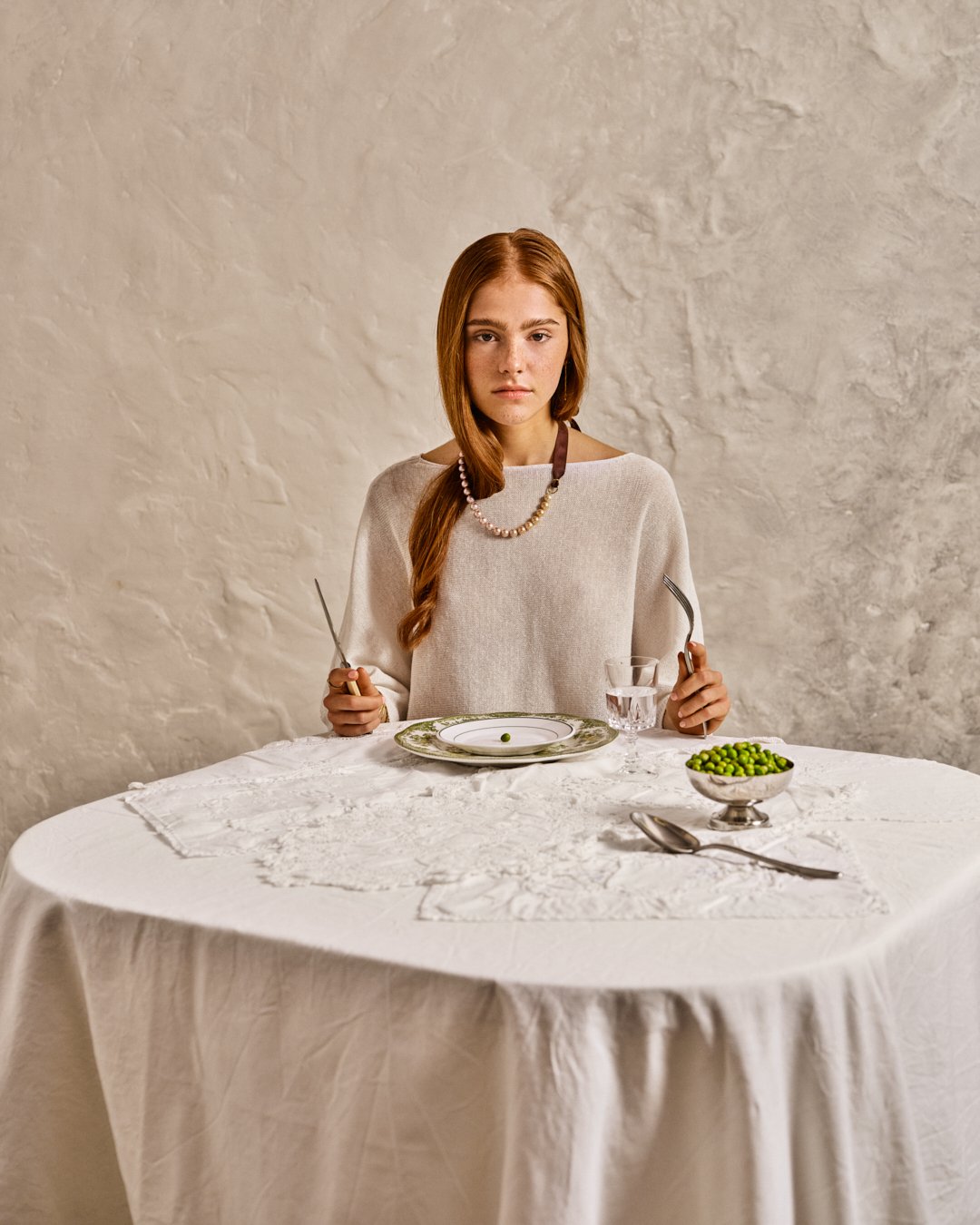 Une jeune femme aux cheveux roux assise à une table avec une assiette, un verre d'eau et un bol de petits pois, dans une ambiance simple et élégante.
