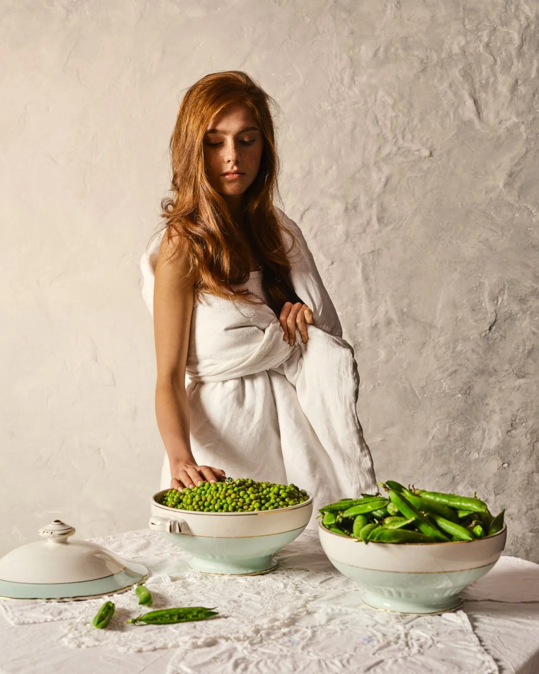 Jeune femme aux cheveux roux, enveloppée dans un tissu blanc, se tient devant une table avec deux grands bols contenant des pois verts et des concombres. La scène est lumineuse, avec un fond blanc texturé.