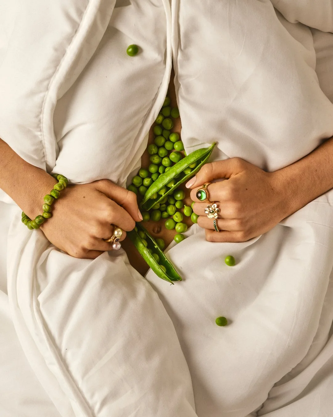 Mains portant des bijoux, entourant une coquille de pois verts ouverte, avec des pois verts tombés dessus, sur un corps habillé en blanc.